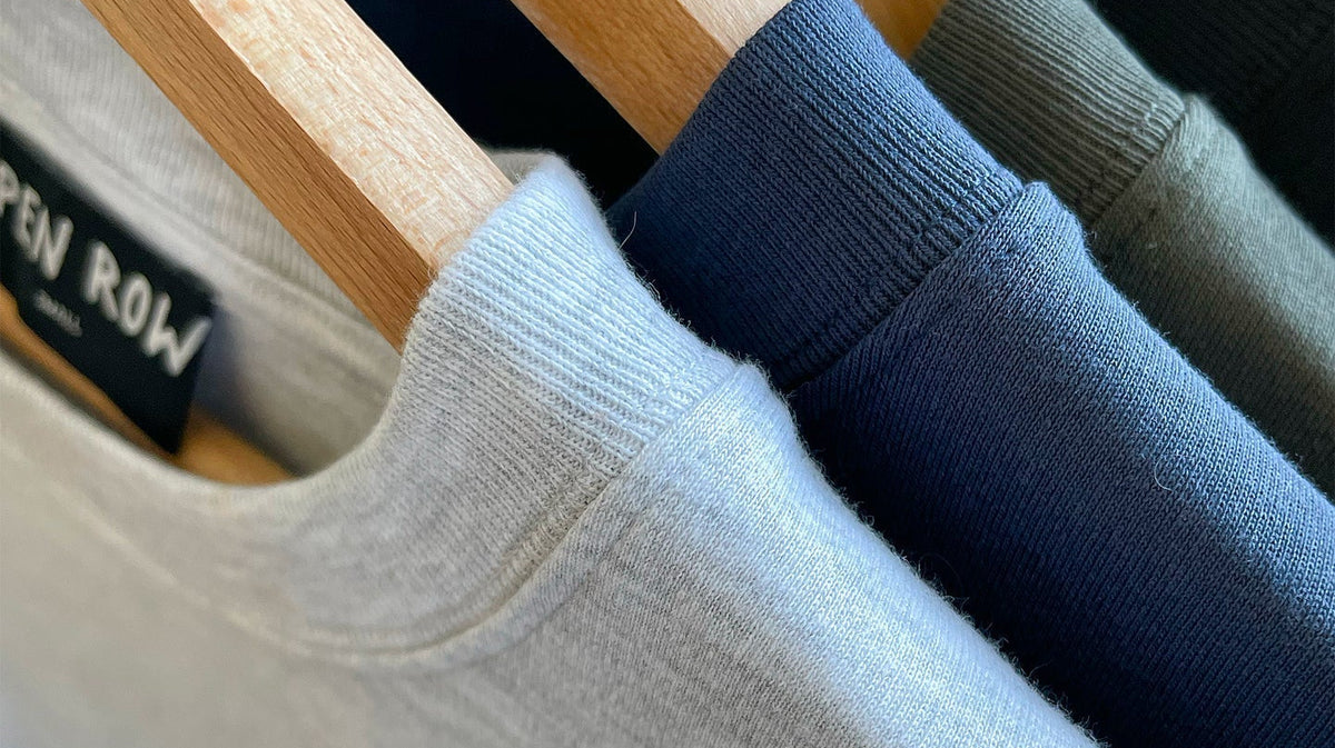 Close-up of Open Heart Tees in gray, navy blue, khaki and black, hanging on wooden hangers. A black neck label reads OPEN ROW.