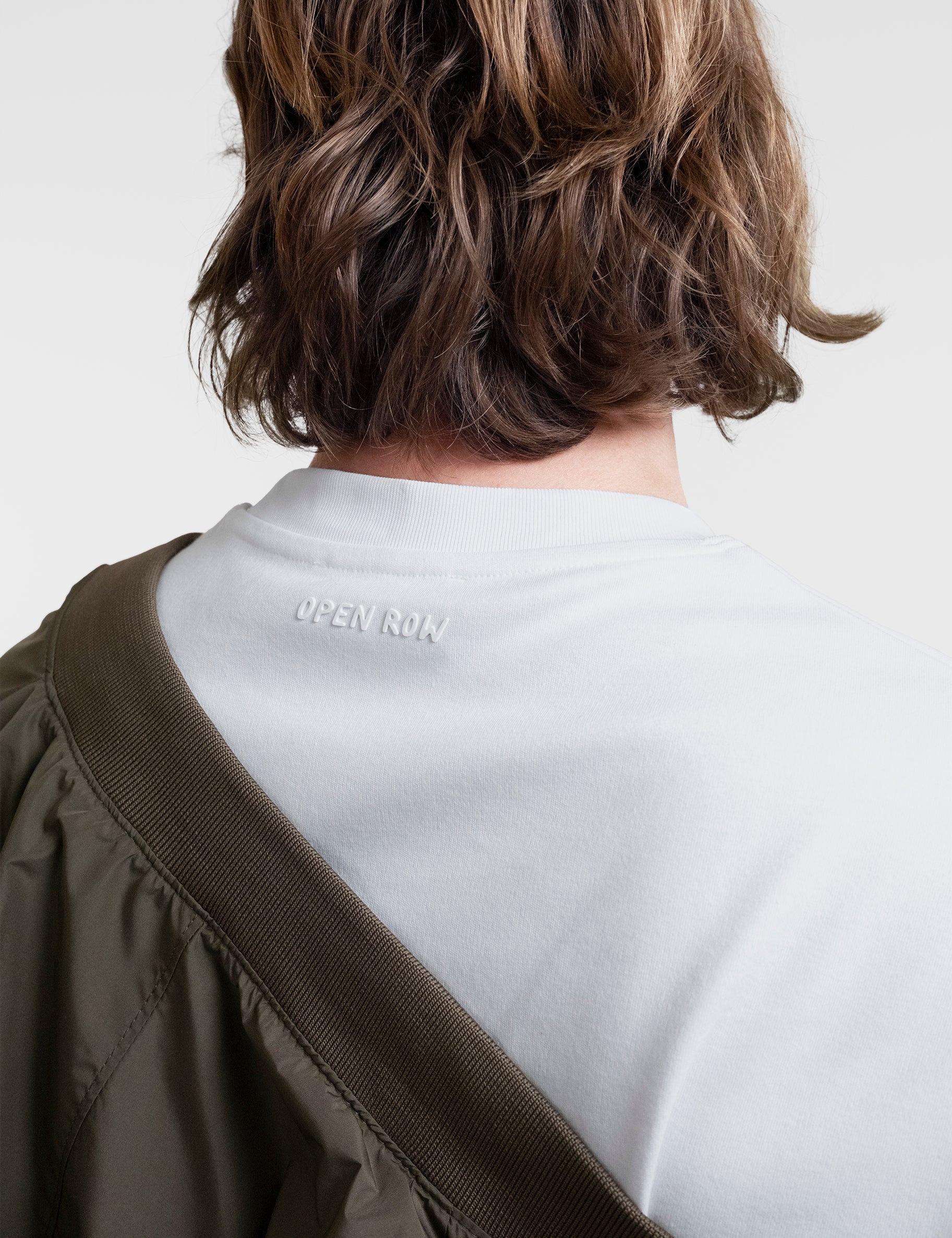 Open Row Men's Tank Tee in white, closeup back view paired with a khaki green jacket, embossed logo detail.