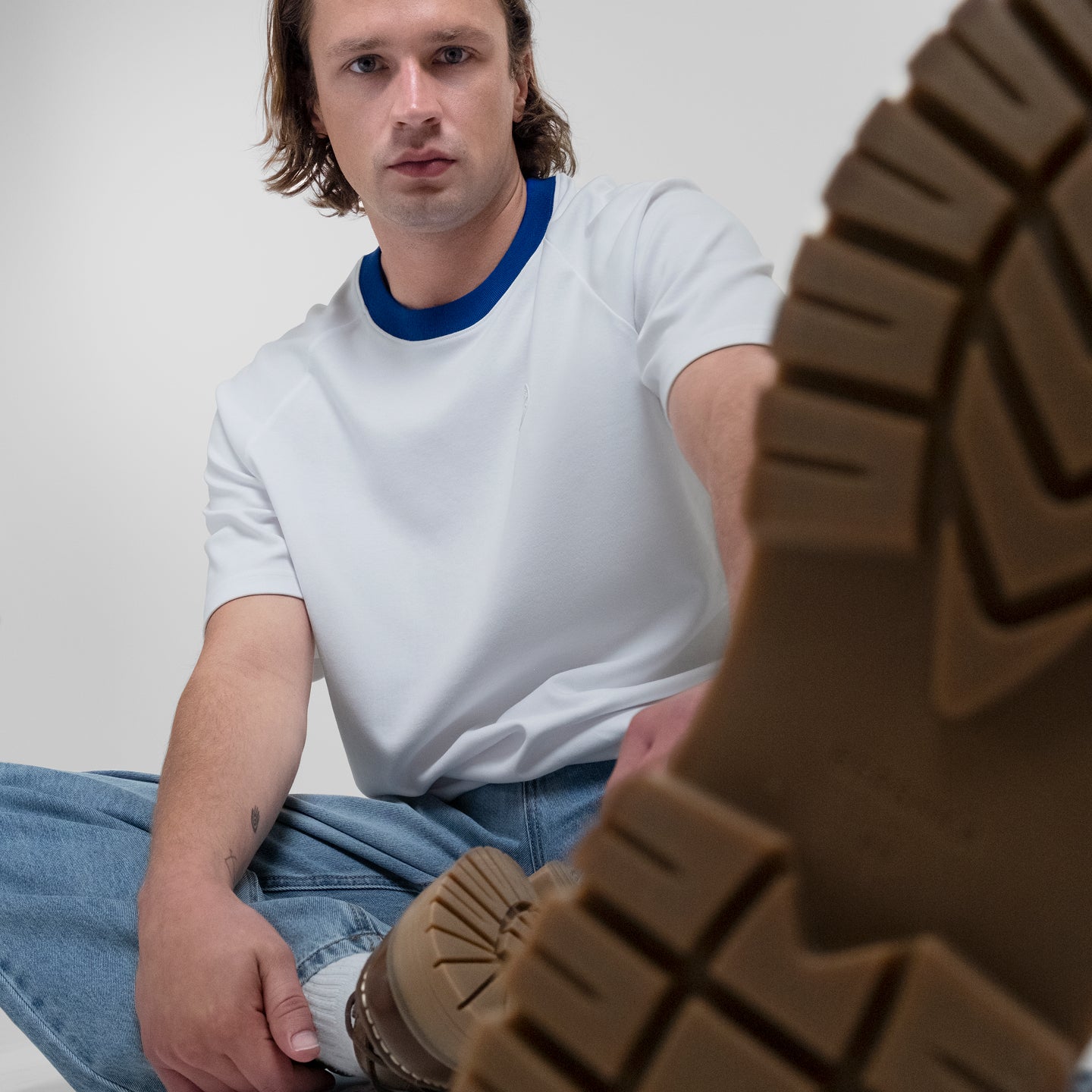A man wearing a white Runaway Tee with a blue collar, sitting cross-legged and holding up a close-up of a brown lug sole.