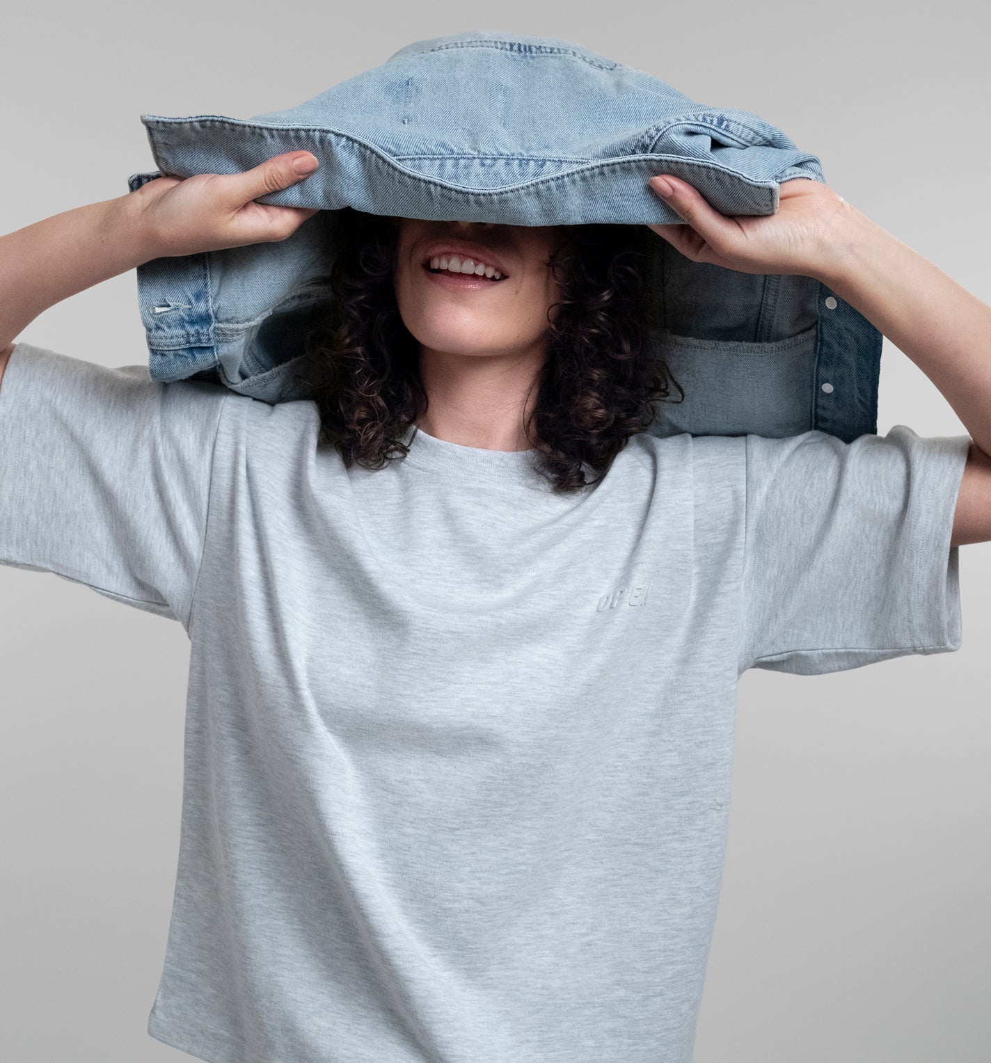 A woman wearing a light grey Open Heart Tee holds a light blue denim jacket over her head like a hat, set against a solid light gray background.