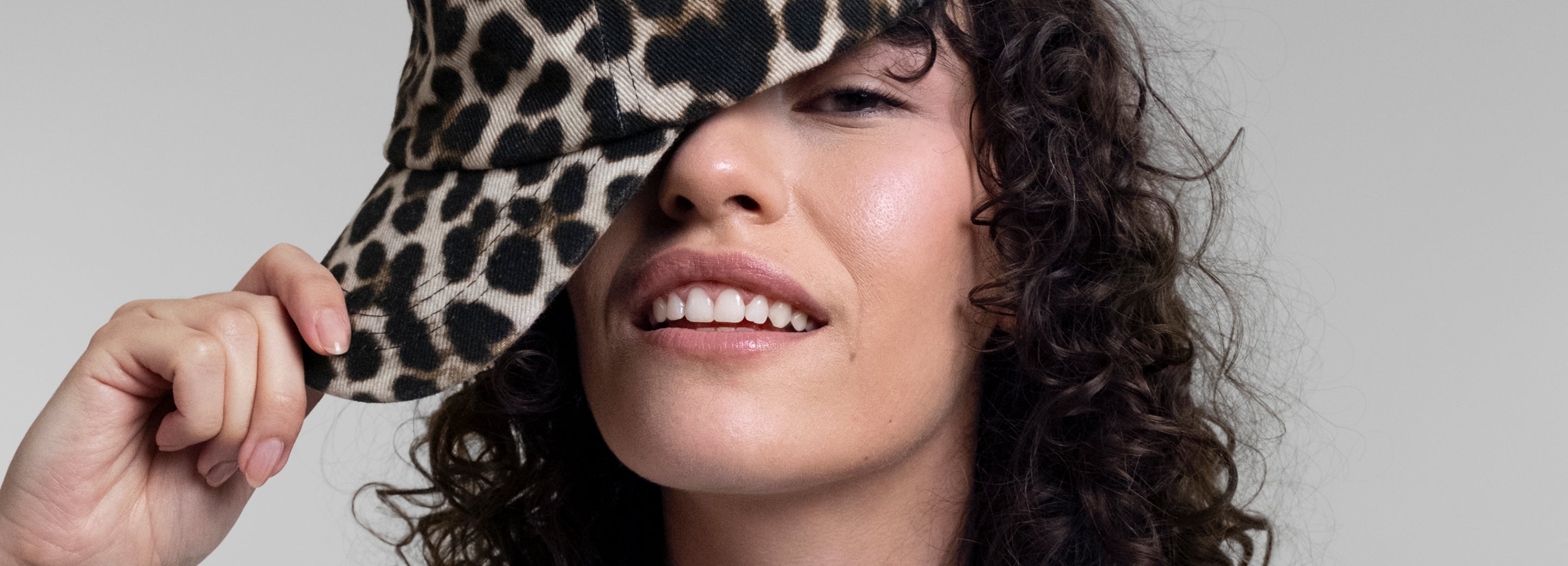A close-up lifestyle shot of a female model with curly dark hair wearing a leopard-print baseball cap, holding it up with her hand against a solid light gray background.