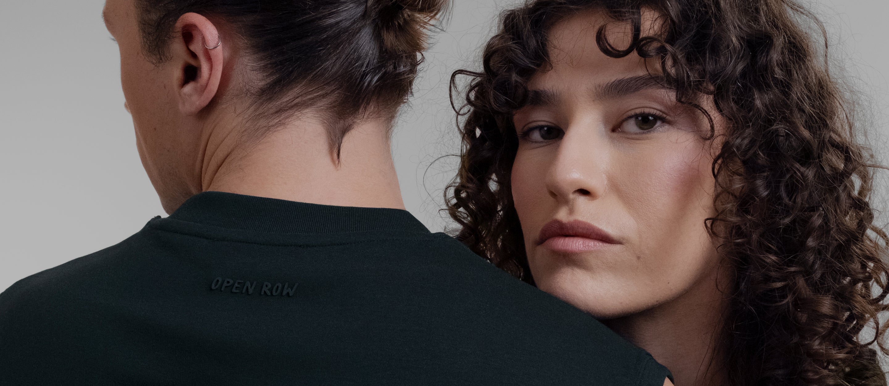 A close-up of a female model with curly brown hair looking over a male model's shoulder, both wearing Open Row Tank Tees with the 'OPEN ROW' logo on the back.