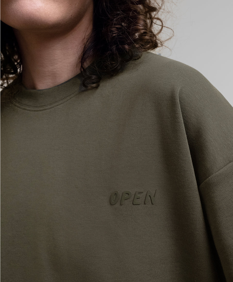 Close-up of a person wearing a khaki Open Heart Tee with the word 'OPEN' embroidered on the heart, against a plain light gray background.