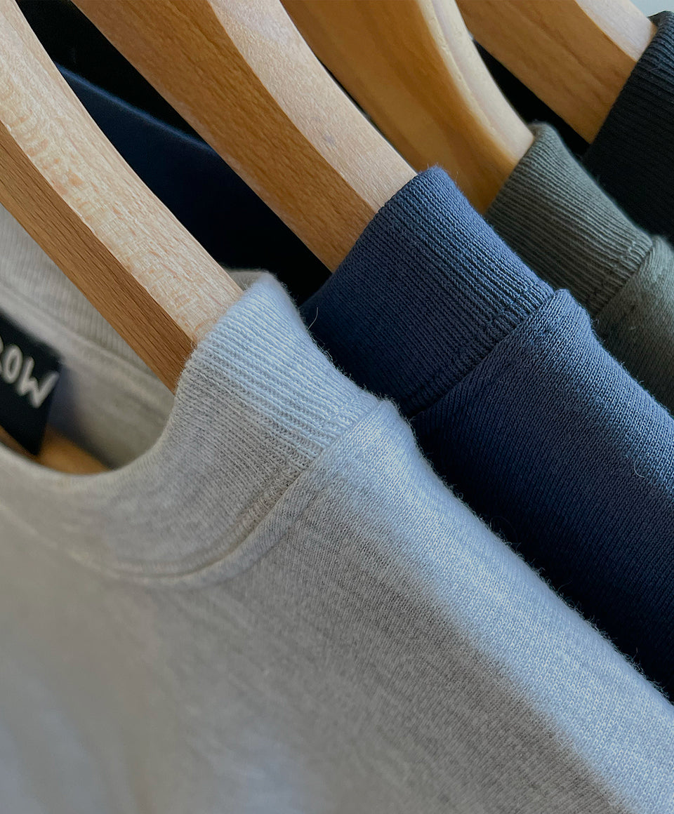 Close-up of Open Heart Tees in gray, navy blue, khaki and black, hanging on wooden hangers. A black neck label reads OPEN ROW.