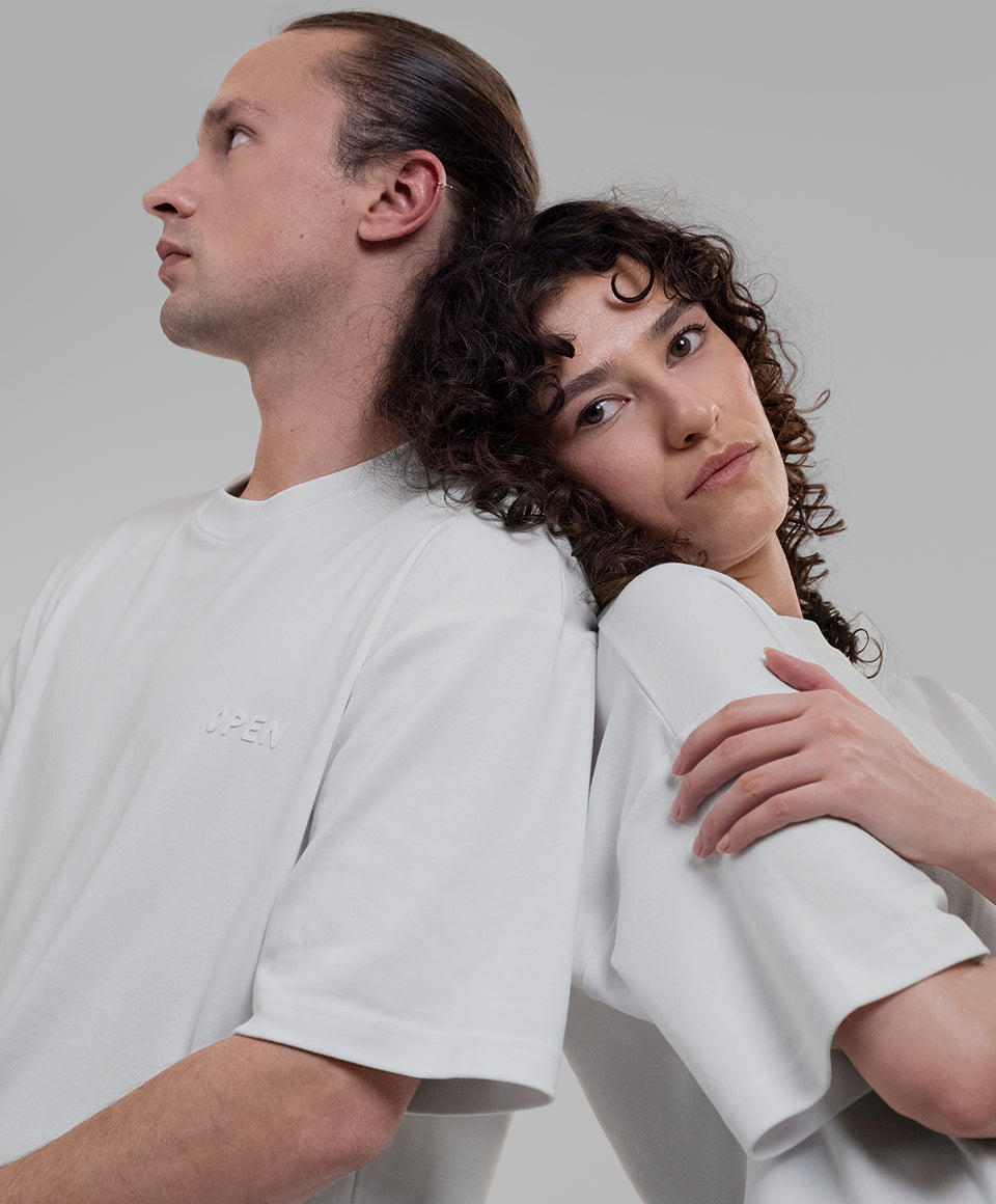 Two models wearing matching white Open Heart Tees with embossed 'OPEN' logos on the heart, posed back-to-back against a solid light gray background.