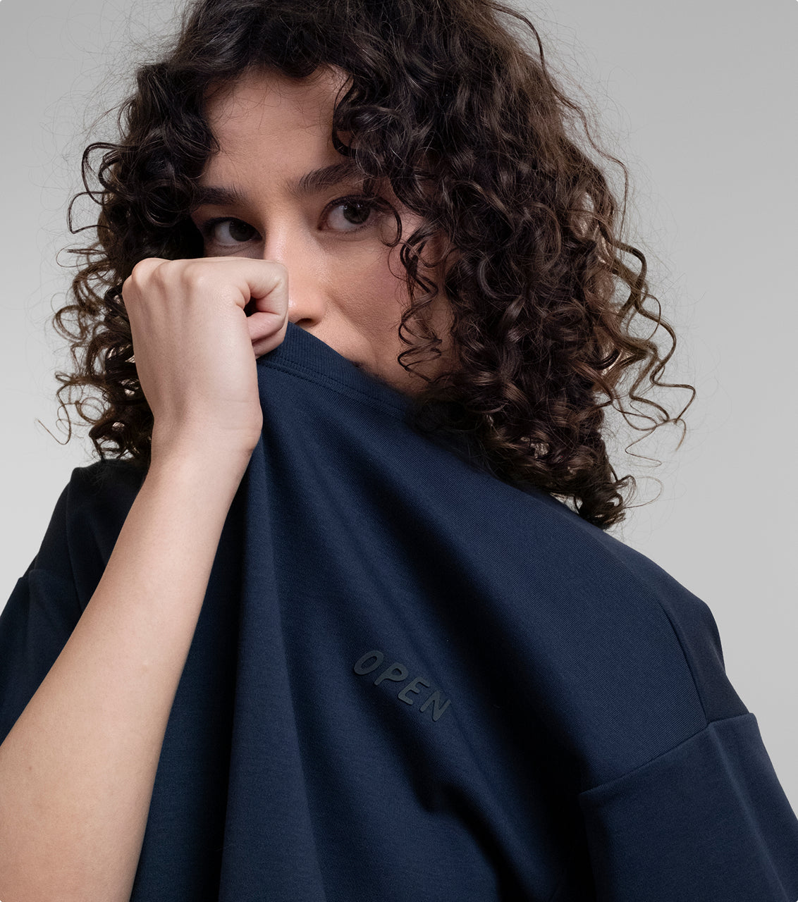 A close-up lifestyle shot of a model with curly brown hair pulling up the collar of a navy Open Heart Tee. The embossed 'OPEN' logo is printed over her heart.