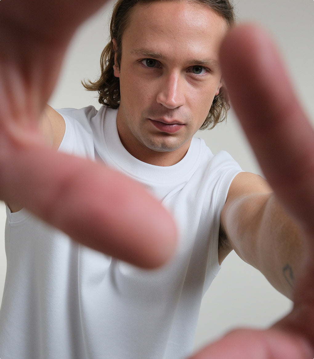 A close-up portrait of a man wearing a white Open Heart Tee, with his hands framing the shot in the foreground against a neutral light gray background.