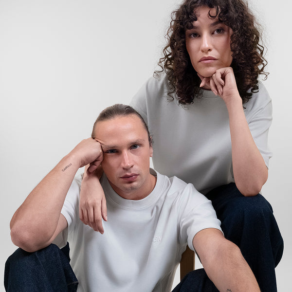 A man and a woman posing together in matching white Open Heart Tees and dark jeans against a plain off-white background.