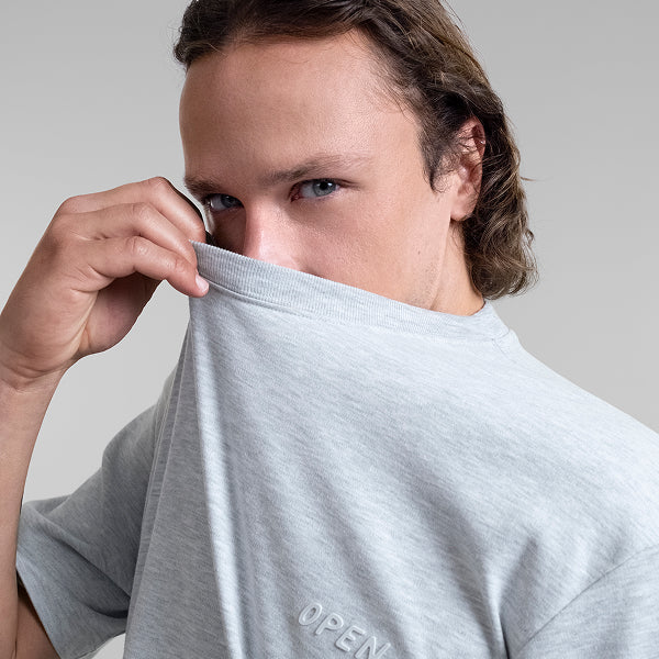 A close-up of a male model wearing a light gray Open Heart Tee, holding the collar up with his hand. The 'OPEN' logo is printed in a matching light grey over his heart.