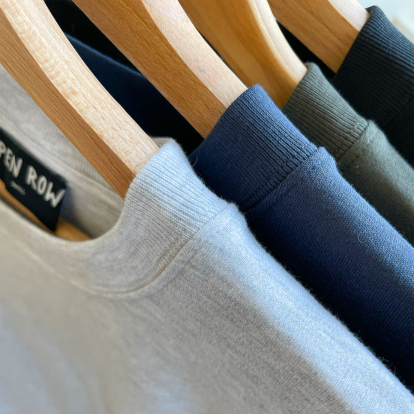 Close-up of Open Heart Tees in grey, navy, khaki and black, hanging on wooden hangers. A black neck label reads OPEN ROW.
