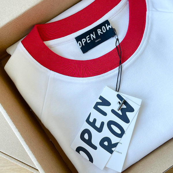 A folded white Runaway Tee with red collar, shown inside a cardboard box. The brand label reads 'OPEN ROW', and a matching paper hangtag is attached.