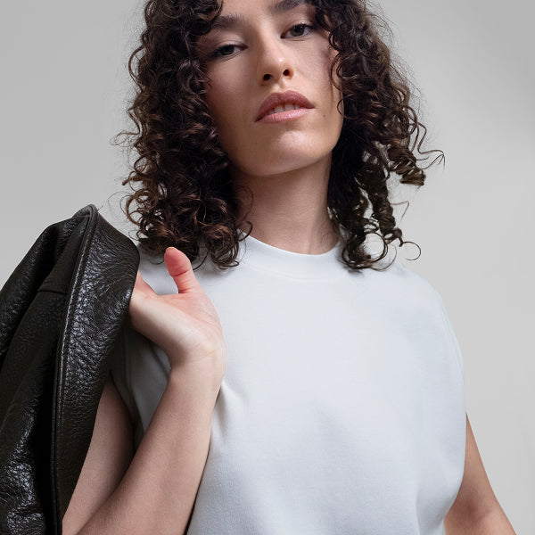A close-up of a woman with curly dark hair wearing a white Open Heart Tee, and holding a black leather jacket against a light gray studio background.
