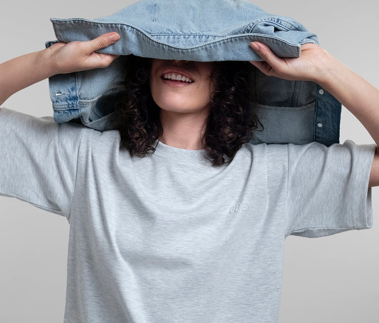 A woman wearing a light grey Open Heart Tee holds a light blue denim jacket over her head like a hat, set against a solid light gray background.
