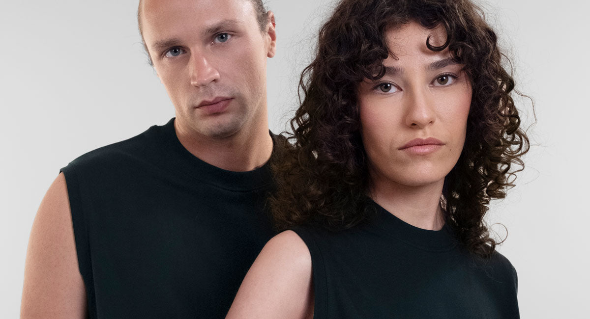 A man and a woman model wearing matching black Open Row Tank Tees, posed against a plain light gray studio background.